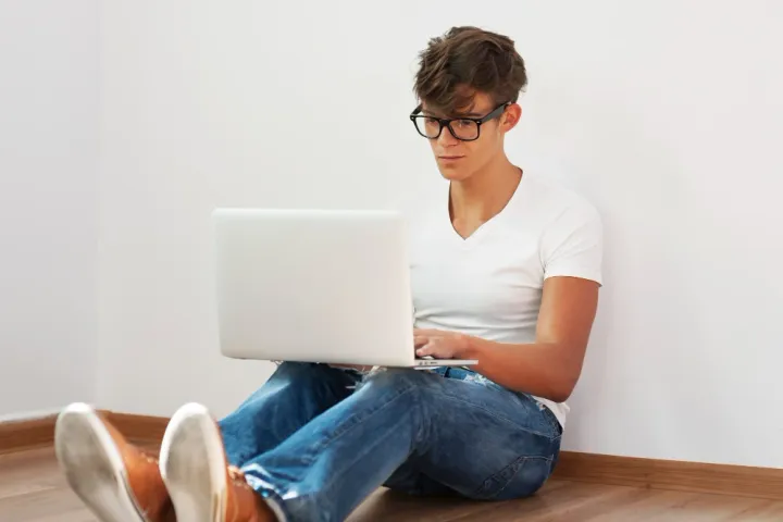 Jovem de cerca de 16 anos, sentado no chão com um laptop apoiado nas pernas, usando óculos e olhando para a tela, parecendo focado.